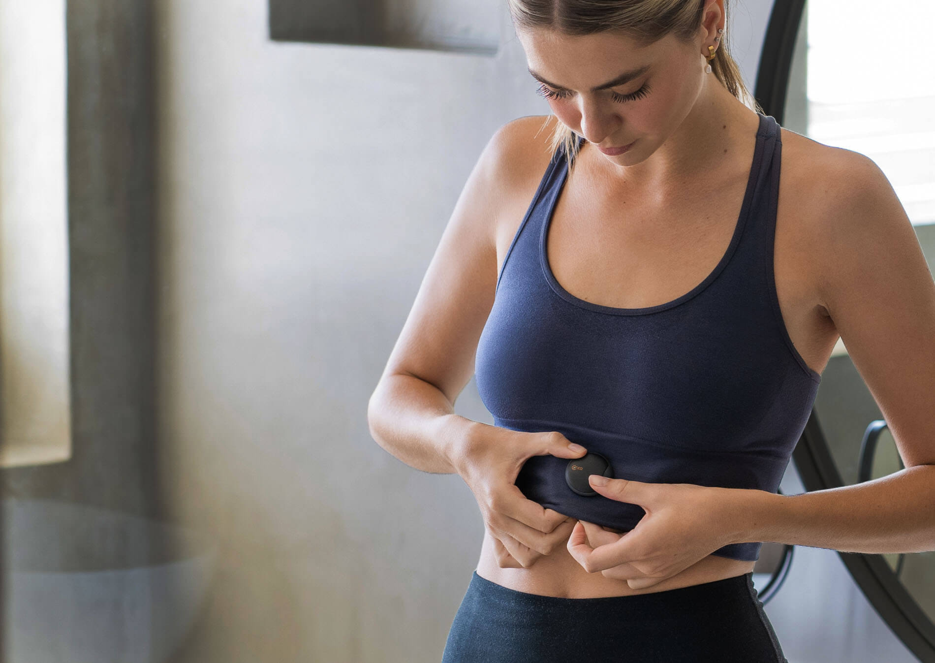 A woman in workout attire attaches the Oxa breathing coach device to her waistband, preparing for a guided breathing exercise to enhance her wellness routine